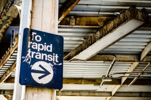 In the foreground, the photo depicts a rusted sign with "To rapid transit" and an arrow. The sign is marked with tagging-style graffiti. In the background are rusted iron girders, part of the infrastructure of the L train.
