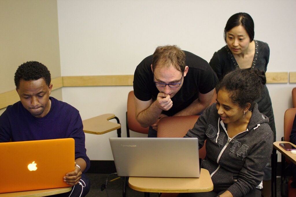 A photo of four people. Two of them are sitting and looking at laptops, while two of them are standing and looking at the laptops thinking. Only one person is smiling.