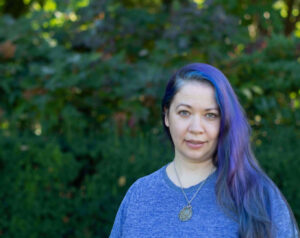 Kaylea Champion (a woman with long blue and purple hair, wearing a blue shirt, on a background with trees)